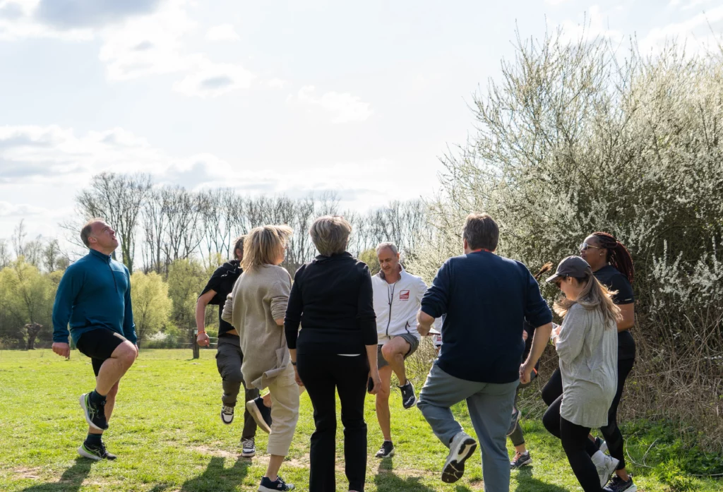 Un groupe de personnes adultes faisant des exercices d’échauffement en cercle dans un parc, en plein air, sous un ciel partiellement nuageux, avec des arbres en arrière-plan. Chacun lève un genou en l'air dans un mouvement coordonné, suggérant une activité sportive collective ou un atelier bien-être.